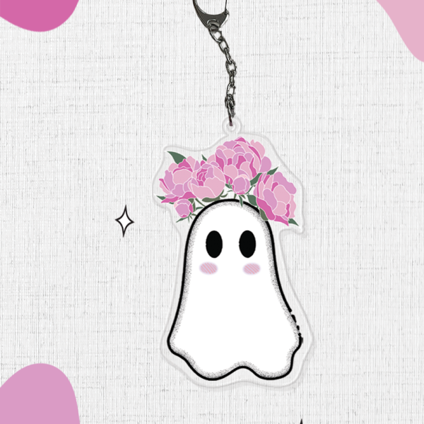 Introducing the Jood Ghost Keychain: a charming accessory showcasing an adorable ghost with rosy cheeks and pink flowers growing from their head. The background boasts abstract pink shapes and scattered sparkles, adding a whimsical and playful touch.