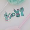 Sticker of four green zombie hands forming the word "LOVE" in American Sign Language, there are two pens framing the sticker, one is pink and one is pastel green.