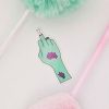 A sticker depicting a green hand with a missing middle finger, revealing bone at the tip and adorned with two purple tattoos, lies on a white surface. Surrounding the sticker are two soft pompom pens, one green and one pink.