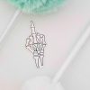 A sticker depicting a skeletal hand making a middle finger gesture is placed between two fluffy pom-pom pens, one green and one white, on a white surface.