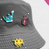 A grey bucket hat adorned with three colorful enamel pins: a blue lock ness monster, a black cartoon mothman with red eyes, and a yellow spaghetti monster. The background features white and pink abstract patterns.