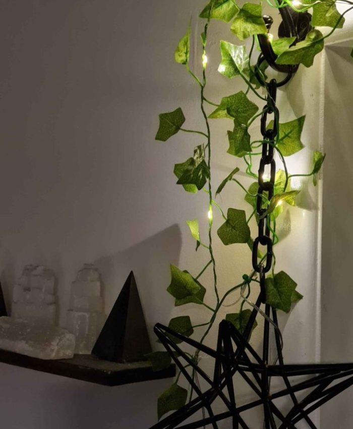 A black geometric star ornament hangs from a chain attached to a wall, adorned with green ivy and small fairy lights. On the shelf below, crystal clusters and pyramids are displayed, creating a mystical and serene ambiance.