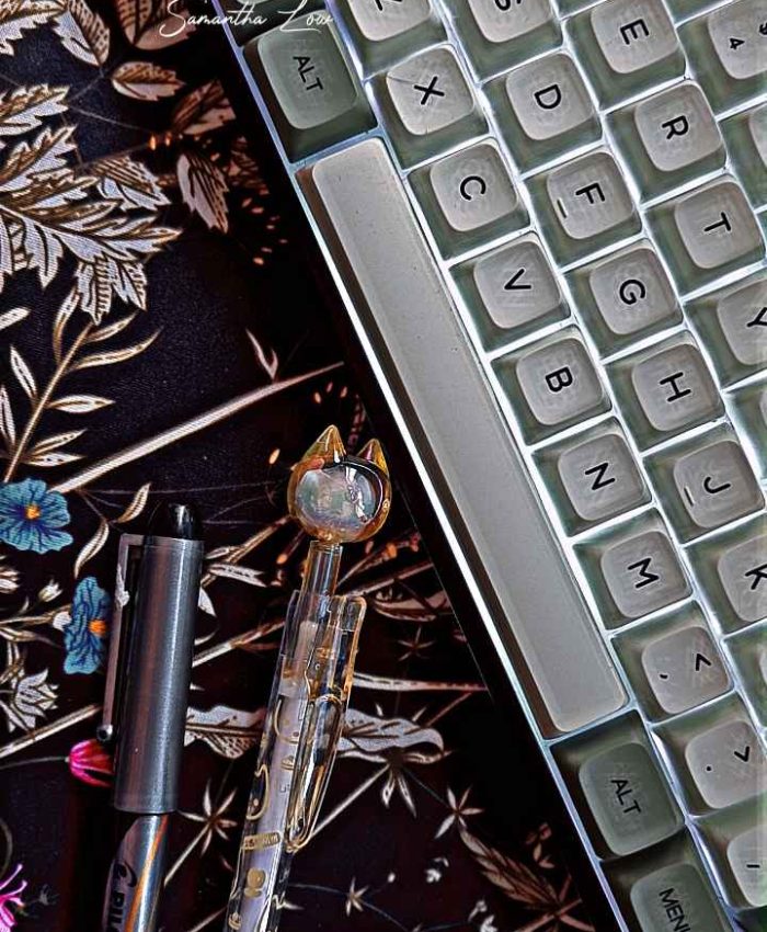 A mechanical keyboard with white and greenish keys is partially visible. Below it lies a black pen and a clear gel pen with a cat-shaped topper. The background is a dark floral-patterned surface.