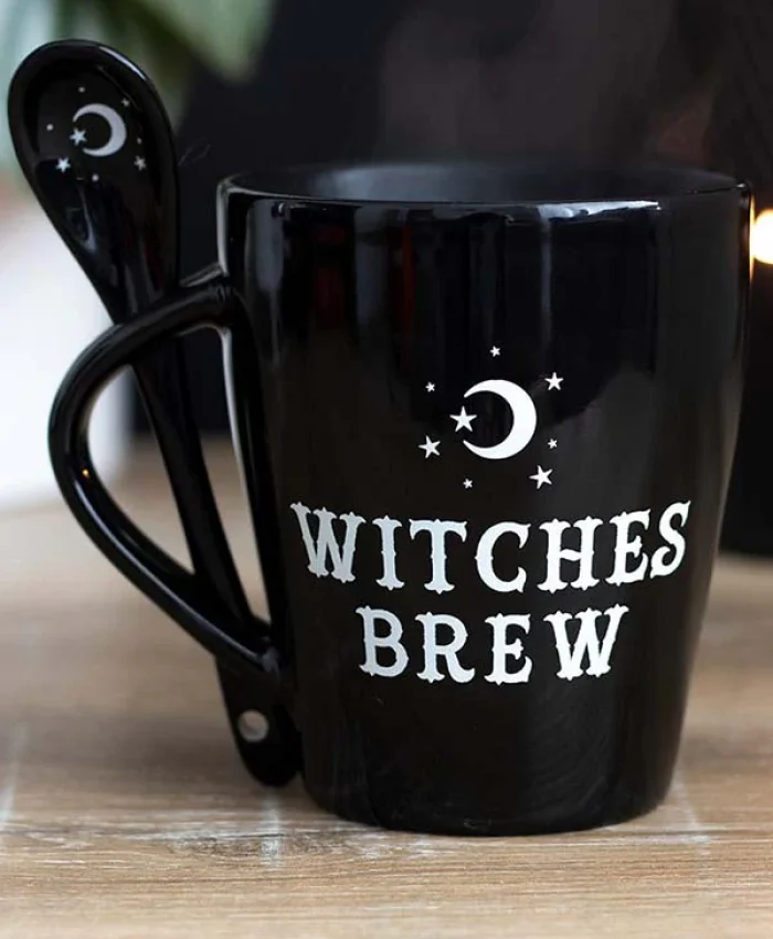 A black coffee mug with white text that reads "Witches Brew" surrounded by stars and a crescent moon. A matching black spoon, which also features a crescent moon, is hooked onto the mug's handle. The mug is placed on a wooden surface.
